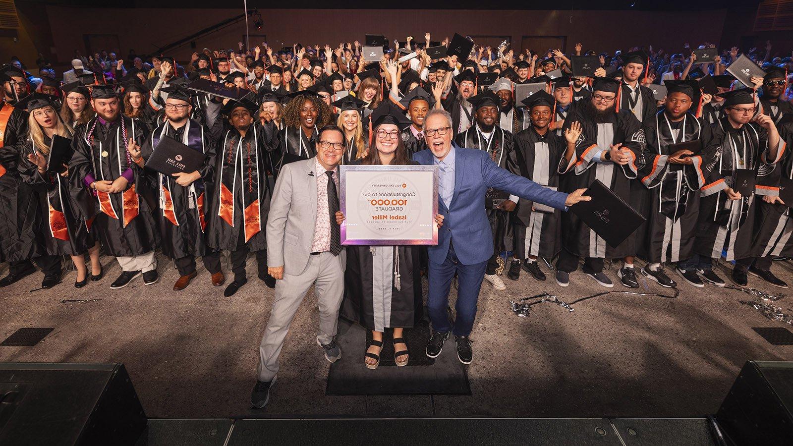 In a large venue full of college graduates, two men in suits stand on opposite sides of a woman in a cap and gown holding a framed photo that reads "满帆大学 - Congratulations to our 100,000毕业 Isabel Miller."