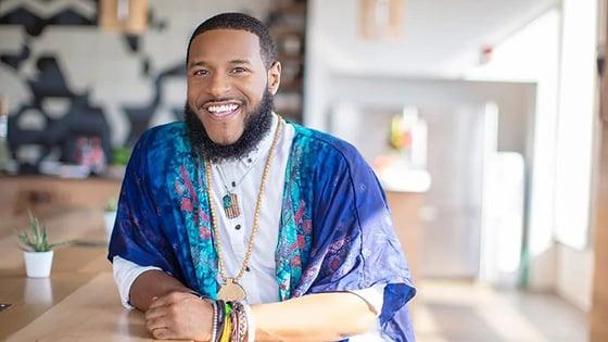 Rico Robinson stands in a café. He is wearing a blue-patterned wrap over a white button-up shirt. 他在微笑.